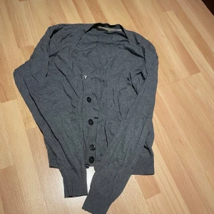 Grå kofta från Massimo Dutti - Stilren grå kofta från Massimo Dutti med knappar framtill. Perfekt för lager-på-lager och har långa ärmar samt ribbade muddar. Den har en defekt då en av knapparna är sönder som man kan se på första bilden. Enkelt att lösa bara att jag inte orkar göra det.