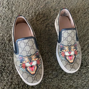 Gucci slip-on sneakers med tigertryck - Gucci slip-on sneakers i beige canvas med klassiskt GG-mönster och färgstarkt tigertryck framtill. Skorna har vita gummisulor, svarta läderdetaljer och grönt/rött band bak. Perfekt statement-piece för dig som vill sticka ut.