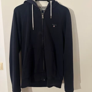 Svart hoodie från Gant med dragkedja - Svart hoodie från Gant med vit snörning i huvan och grått innerfoder. Tröjan har dragkedja framtill, två fickor och diskret Gant-logga på bröstet. Perfekt för en chill och stilren look. En liten Fläck bara går bort i tvätten 