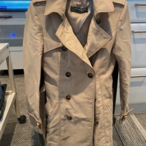 Beige trenchcoat från Zara Woman - Stilren beige trenchcoat från Zara Woman med klassisk dubbelknäppning, axelklaffar och bälte i midjan. Jackan har långa ärmar med spännen vid handlederna och snygga detaljer som ger en tidlös look. Perfekt för dig som vill ha en clean och trendig stil.