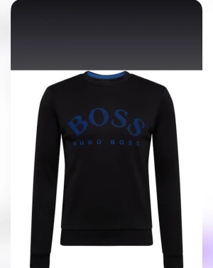 Svart sweatshirt från Hugo Boss - Svart sweatshirt från Hugo Boss med blått BOSS-tryck på bröstet. Tröjan har rund halsringning, långa ärmar och ribbade muddar vid ärmslut och nederkant. Perfekt för en stilren och avslappnad look.