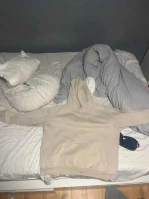 Beige Essentials Fear of God hoodie - Säljer en beige hoodie från Essentials Fear of God med tryck på bröstet och ärmen. Tröjan har huva, stor magficka och ribbade muddar. Perfekt för dig som gillar clean streetwear och vill ha en snygg, enkel look. Materialet är mjukt och känns riktigt skönt mot huden.