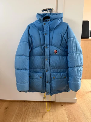 Blå Fjällräven pufferjacka XXXS - Blå pufferjacka från Fjällräven med huva och gula snören. Jackan har tryckknappar, dragkedja och två stora fickor framtill. Ikonisk röd Fjällräven-logga på bröstet. Perfekt för kalla dagar!                          Den har hål som sytts samt lite solblekt (bild på det största defekterna)  Därav priset  Passar mig som brukar ha xs/s i jackor  Bara skriva om ni har frågor 🩷