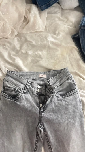 Grå bootcut jeans från Nelly - grå jeans från Nelly med bootcut passform. Jeansen har svarta broderade detaljer på bakfickorna, klassisk femficksdesign och dragkedja med dubbla knappar framtill