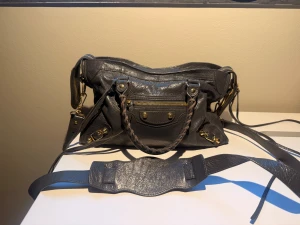 Balenciaga svart axelväska i skinn - Säljer en svart axelväska från Balenciaga i mjukt skinn med flätade handtag, guldfärgade detaljer och dragkedja framtill. Väskan har flera fack och en bred axelrem för extra komfort. Tillhörande spegel finns, den har tyvärr en defekt. Kan skicka bild om det behövs!