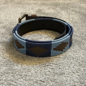 Liknande pampeano bälte - Snyggt bälte som liknar pampeano med geometriskt mönster i ljusblått, mörkblått och brunt. Finns inte kvar i lager. Bältet har en klassisk mässingsfärgad spänne och är gjort i skinn med broderade detaljer. Perfekt för att ge outfiten en cool touch. Helt nytt aldrig använt. Från Nordfors.