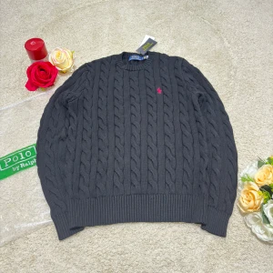 Mörkgrå kabelstickad tröja Polo Ralph Lauren - Snygg mörkgrå kabelstickad tröja från Polo Ralph Lauren med klassisk rund hals och röd broderad logga på bröstet. Tröjan har långa ärmar och ribbade muddar vid ärmslut och nederkant. Perfekt för dig som gillar stilren och tidlös design.