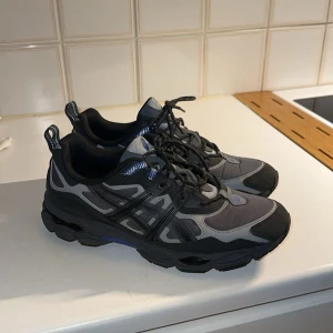 ASICS Gel  - Säljer ett par ASICS Gel sneakers i svart och grå med blå detaljer. Skorna har en chunky sula, snörning och reflekterande detaljer baktill. Perfekta för dig som gillar sportig stil och vill ha bekväma skor till vardags. Materialet är syntet och textil.