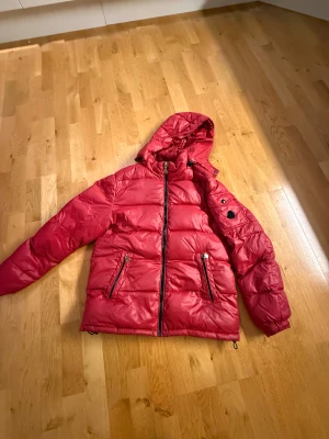 Röd pufferjacka med huva - Röd pufferjacka med huva och två framfickor med dragkedja. Jackan har quiltad design, lång ärm och en ficka med knapp på ena armen. Materialet är glansigt och syntetiskt, perfekt för kalla dagar. Dragkedja framtill och elastiska muddar vid ärmslut.