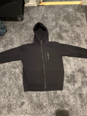 Svart hoodie från Sail Racing M - Svart hoodie från Sail Racing i storlek M. Tröjan har hel dragkedja framtill, huva med snörning och en bröstficka med dragkedja. Insidan är fodrad med mjuk fleece för extra komfort. Snygg logga på bröstet och klassisk passform. Den är bara använd 4 gånger då den var förstor! Nyskick!