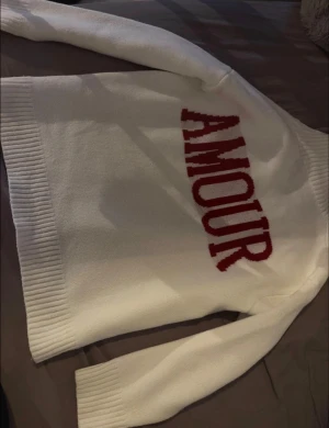 AMOUR - Vit med bred ribbad krage och muddar. På ryggen står det AMOUR i stora röda bokstäver. Tröjan är stickad och har en loose fit med långa ärmar. Perfekt för dig som gillar statement-plagg och vill ha något mysigt men ändå snyggt.