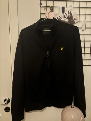 Svart ziptröja från Lyle & Scott - Svart långärmad ziptröja från Lyle & Scott med gul broderad logga på bröstet. Tröjan har en half zip framtill och är tillverkad i mjuk bomull. Perfekt för dig som gillar stilrena och klassiska plagg med en sportig touch.