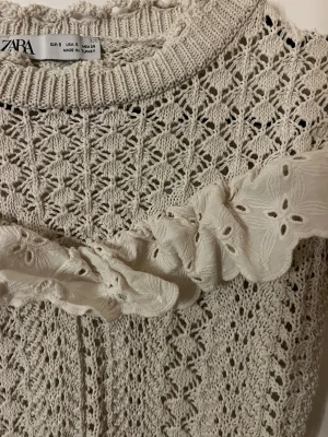 Beige stickad topp med volang från Zara - Supersöt beige stickad topp från Zara med korta ärmar och rund hals. Toppen har ett detaljerat hålmönster och en bred volang med broderade blommor över bröstet. Perfekt för dig som gillar romantiska och unika plagg med mycket detaljer.