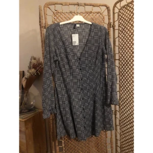 Rutig, A-linjeformadklänning från H&M - Snygg rutig klänning från H&M i svart och vit med v-ringning och knappar framtill. Klänningen har långa ärmar och en kort, lätt utställd passform som ger en chill vibe. Klänningen är oanvänd, och 46 i storlek (men upplevs mindre i storlek).