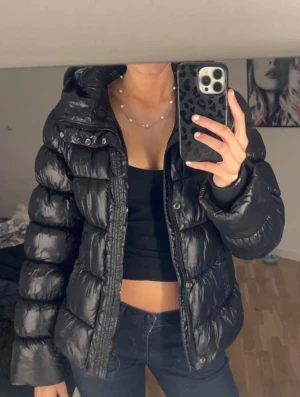 Svart pufferjacka från Superdry - Säljer en svart, glansig pufferjacka från Superdry med huva och dragkedja framtill. Jackan har quiltad design, två fickor med knappstängning och en cool, croppad passform som ger en trendig look. Perfekt för kalla dagar och snygg att styla med jeans.