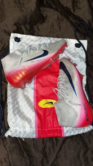 Nike Mercurial Zoom fotbollsskor - Nike Mercurial Zoom fotbollsskor i ljusgrått med snygga mörkblå swoosh-detaljer och en röd till rosa gradient på sulan. Skorna har snörning, platt sula och är tillverkade i syntetmaterial med mesh för extra ventilation. Perfekt för dig som vill sticka ut på planen.