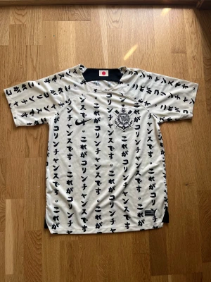 Corinthians fotbollströja (sällsynt) - Unik vit fotbollströja från Corinthians med svarta japanska tecken över hela tyget. Framsidan har klubbens emblem och Nike-logga, baksidan har 'Paulinho' och nummer 15 i svart med guldkanter. Tillverkad i lätt polyester, kortärmad och med rund hals. Storlek M.