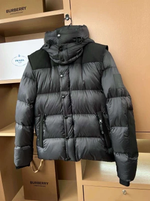 Svart pufferjacka från Burberry - Svart pufferjacka från Burberry med avtagbar huva och snygga detaljer. Jackan har stora, quiltade sektioner, dragkedja och knappar framtill samt två fickor med dragkedja. Burberry-logga på ärmen och justerbar snörning vid huvan. Perfekt för kalla dagar.