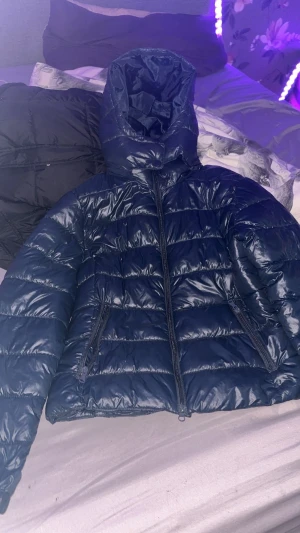 Mörkblå pufferjacka med huva - Säljer en mörkblå pufferjacka med glansig yta och huva. Jackan har två sidofickor med dragkedja och quiltad design. Perfekt för kalla dagar och har en snygg, modern look som funkar till streetwear. Storlek M men passar s oxå 