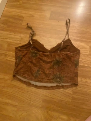 Brun blommig mesh linnetopp - Säljer en brun mesh linnetopp med blommigt mönster och tunna justerbara axelband. Toppen har en lätt croppad passform och spetsdetaljer längs bysten. Perfekt för dig som gillar vintagevibbar och unika detaljer.