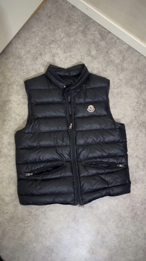 Svart Moncler dunväst med dragkedja - Svart dunväst från Moncler med hög krage och två fickor med dragkedja. Västen har ett quiltat mönster och Moncler-logga på bröstet. Materialet är glansig polyester och dunfyllning, perfekt för lager-på-lager under höst och vinter. OBS! Den har två små hål bara