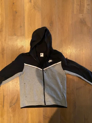 Svart och grå vindjacka från Nike - Niketech med huva i svart och grått, med vit detalj över bröstet. Jackan har dragkedja framtill och två sidofickor. Snygg sportig look med klassisk Nike-logga på bröstet. Perfekt för dig som gillar streetwear och sport.