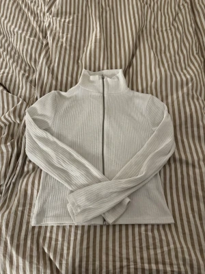 Vit ribbad zip-kofta  - Vit ribbad kofta med hel dragkedja och krage. Tröjan har långärmad passform, är figurnära, och  är lite tunn. Storlek M från Lager 157