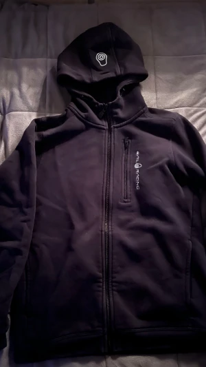 Svart hoodie från Sail Racing med dragkedja - Svart hoodie från Sail Racing med hel dragkedja och broderad logga på bröstet samt huvan. Tröjan har en mjuk insida, huva med vita detaljer och en praktisk bröstficka med dragkedja. Perfekt för dig som gillar sportig och clean stil.