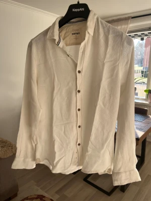 Vit slim fit skjorta från Koton - Vit skjorta från Koton i slim fit med snygga bruna knappar och klassisk krage. Materialet är lätt och luftigt bomull, perfekt för varmare dagar. Skjortan har långa ärmar med förstärkta armbågslappar och en stilren, enkel design som funkar till många looks.