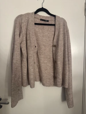 Beige stickad kofta från 157, S - Mysig beige stickad kofta från 157 i storlek S. Koftan har långärmade ärmar, ribbade muddar och knappar framtill. Perfekt för lager-på-lager och chill dagar. Enkel och stilren design som funkar till många outfits.