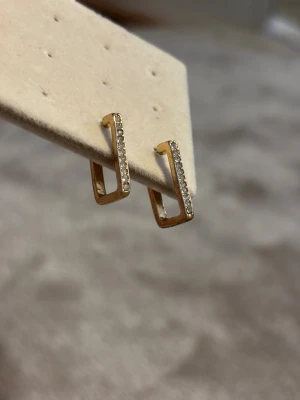 Guldiga örhängen med stenar - Säljer ett par stilrena örhängen i guldtonad metall med rektangulär form. Framsidan är dekorerad med små gnistrande stenar som ger extra bling. Perfekt för dig som gillar minimalistisk design med en touch av glamour. 