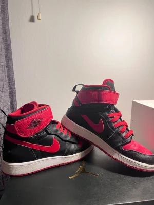 Nike Air Jordan 1 Hi FlyEase röd/svart - Nike Air Jordan 1 Hi FlyEase sneakers i svart och rött med klassisk Jordan-logga på ankeln. Skorna har snörning, kardborreband och dragkedja för enkel påtagning. Tillverkade i skinn med vit och röd yttersula. Snygg high-top siluett som sticker ut. NÅTT MÄRKE PÅ SKON!!