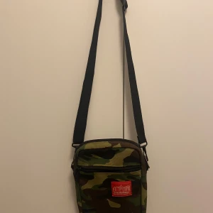 Axelväska Manhattan Portage i camouflage - Snygg axelväska i kamouflage / militärmönster från Manhattan Portage. Väskan har en justerbar axelrem och en dragkedja. Köpt för några år sedan, sparsamt använd. 