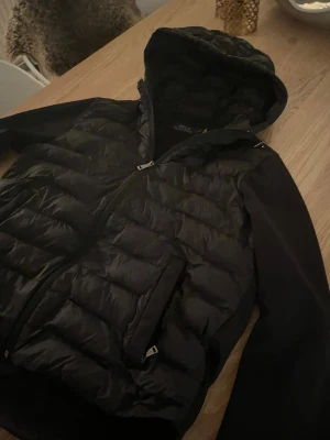 Svart dunjacka från Polo Ralph Lauren - Svart dunjacka från Polo Ralph Lauren med huva och quiltad front. Jackan har långa ärmar i slät polyester, dragkedja framtill och två sidofickor med dragkedja. Klassisk Polo-logga broderad på bröstet. Perfekt för kyliga dagar. Pris kan diskuteras vid snabb affär 