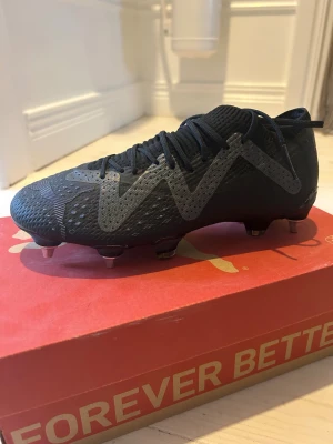 Puma Future Ultimate fotbollsskor svart - Säljer ett par svarta Puma Future Ultimate fotbollsskor i storlek 36. Skorna har snörning, platt sula med metall-dobbar och en cool grafisk detalj i grått på sidan. Ovandelen är tillverkad i syntetmaterial med textur för extra grepp och kontroll på planen.