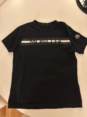 Svart Moncler t-shirt med logga - Svart t-shirt från Moncler med vit rand och stor broderad logga över bröstet. Klassisk rund hals och korta ärmar. Moncler-märke på vänster ärm ger extra premiumkänsla. Perfekt för dig som gillar stilrena designer med tydlig branding.