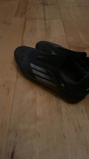 Adidas F50 svarta fotbollsskor - Svarta Adidas F50 fotbollsskor med silvriga detaljer och smal, aerodynamisk form. Skorna har en låg profil och är tillverkade i syntetmaterial för lätthet och snabbhet på planen. Diskret brasiliansk flagga på sidan och F50-logga på ovansidan.