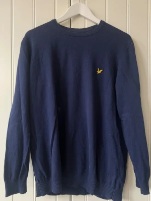 Marinblå stickad tröja Lyle & Scott - Snygg marinblå stickad tröja från Lyle & Scott i storlek XXL (sitter som L). Tröjan har rund hals, långa ärmar och klassisk gul fågel-logga broderad på bröstet. Mjuk och skön kvalitet som passar perfekt till jeans eller chinos. Ribbad kant vid ärmslut och nederkant.
