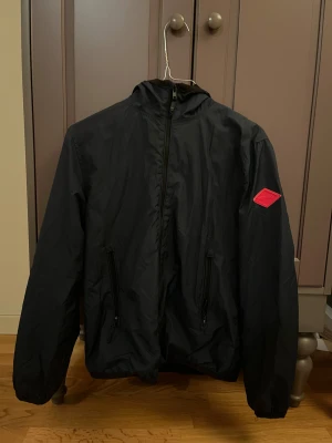 Mörkblå vindjacka från Replay - Mörkblå  vindjacka från Replay med huva och hel dragkedja framtill. Jackan har två fickor med dragkedja och ett rött Replay-märke på ärmen. Tillverkad i lätt polyester med meshfoder för extra komfort. Perfekt för dig som gillar stilren och sportig look.