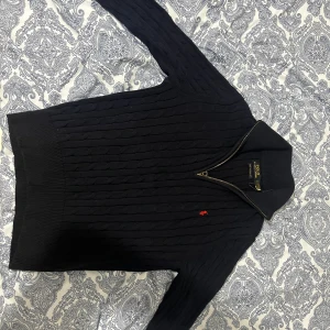 Svart stickad half zip tröja Polo Ralph Lauren - Navy blue halv zip tröja från Polo Ralph Lauren med half zip och kabelstickat mönster. Liten röd logga broderad på bröstet. Hög krage och långa ärmar, perfekt för kyliga dagar. Snygg och klassisk design som funkar till jeans eller chinos.