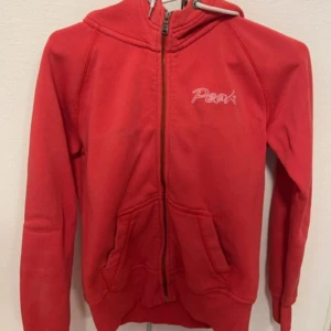  hoodie från Peak Performance  - Rosa/korall färgad hoodie från Peak Performance med dragkedja framtill, broderad logga på bröstet och huva med broderad P. Tillverkad i 80% bomull och 20% polyester. Två fickor framtill och ribbade muddar vid ärmslut och nederkant.använd någon enstaka gång 