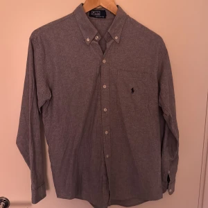Grå skjorta från Polo Ralph Lauren - Klassisk grå skjorta från Polo Ralph Lauren i bomull. Skjortan har button-down krage, bröstficka med broderad logga och knäppning framtill. Långärmad och med en clean, tidlös look som funkar till allt. KAN GÅ NER I PRIS VID EN SNABB AFFÄR!!