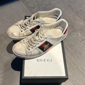 Gucci Ace sneakers med bi-detalj - Gucci Ace sneakers i vitt skinn med klassiska gröna och röda ränder på sidan och broderad guldfärgad bi. Baksidan har metallicröd och metallicgrön detalj. Snygg och ikonisk modell med snörning och platt sula. Kommer med originalkartong.