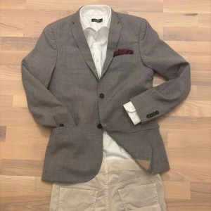 Grå kavaj J.Lindeberg i ull slim fit - Snygg grå kavaj från J.Lindeberg, sydd i ett exklusivt tyg från Angelico – vävt i Biella, Italien. Slim fit-modell i 100% virgin wool, med två knappar framtill, klassiska slag samt beige innerfoder.   Storlek 46R 	•	Axel till axel: 43 cm 	•	Armhåla till armhåla: 48 cm 	•	Armlängd: 63 cm 	•	Längd: 72 cm