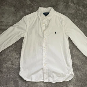 Vit skjorta från Polo Ralph Lauren - Säljer min son klassiska vita skjorta från Polo Ralph Lauren i slim fit-modell. Skjortan har långa ärmar, knappar framtill och den ikoniska broderade loggan på bröstet. Tillverkad i mjuk bomull och passar perfekt till en clean och stilren look.