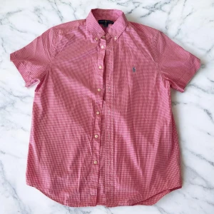 Rutig rosa kortärmad skjorta Ralph Lauren - Kortärmad skjorta från Ralph Lauren i rosa och vitt rutigt mönster. Klassisk button-down krage, knappar framtill och broderad blå logga på bröstet. Skjortan är tillverkad i mjuk bomull och har en avslappnad passform. I jätte fin skick, använd ett par gånger. 