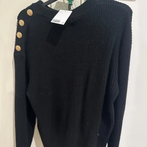 Svart stickad tröja med guldknappar - Svart ribbstickad tröja med rund hals och fyra snygga guldknappar längs ena axeln. Tröjan har lång ärm och en klassisk, lite loose passform. Perfekt för dig som gillar stilrena detaljer och vill ha något som sticker ut lite extra.