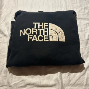 Svart hoodie från The North Face - Svart hoodie från The North Face med stor vit logga på bröstet. Klassisk känguruficka framtill och huva. Mjuk insida och ribbade muddar vid ärmslut och nederkant. Perfekt för chill dagar eller när du vill ha en enkel streetstyle.