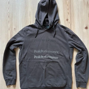 Grå hoodie från Peak Performance - M - Säljer en grå hoodie från Peak Performance med tryckt logga i olika storlekar på bröstet. Tröjan har huva med snörning, känguruficka och långa ärmar. Perfekt för dig som gillar en avslappnad och stilren look.