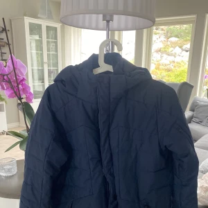 Mörkblå pufferjacka med huva - Säljer en mörkblå pufferjacka med huva och dragkedja framtill. Jackan har två fickor med dragkedja och quiltad design. Perfekt för kalla dagar och riktigt skön att bära. Passformen är normal och jackan är tillverkad i syntetmaterial.